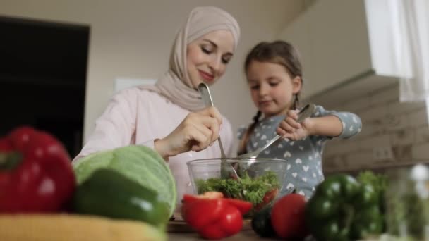 En la moderna y acogedora cocina doméstica pequeña hija adorable ayuda a la joven madre musulmana a cocinar una cena saludable — Vídeo de stock