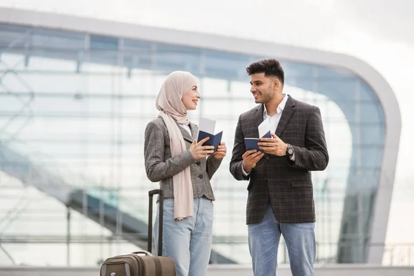 Pareja musulmana con maleta, parada al aire libre en el aeropuerto y revisando su horario de vuelo —  Fotos de Stock