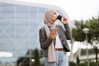 Başörtülü Arap kadın kahve ve telefonla sokakta yürüyor.