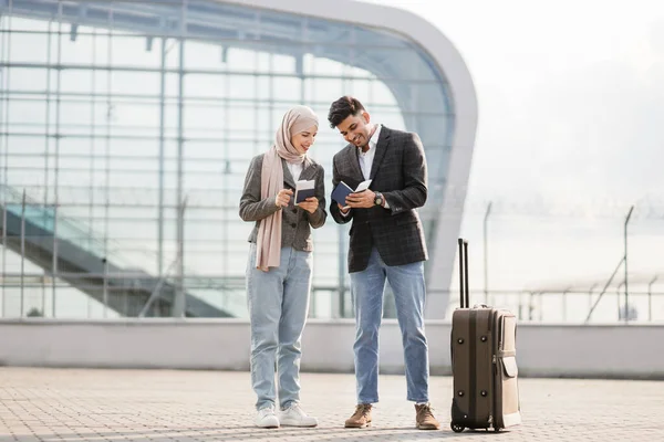 Dama musulmana en hiyab, y hombre árabe, llevando maleta fuera del aeropuerto, sosteniendo pasaportes y billetes —  Fotos de Stock
