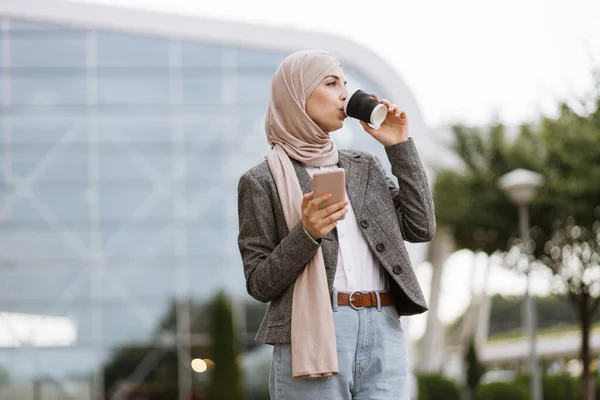 Arab nő fejkendőben sétál az utcán kávéval és telefonnal. — Stock Fotó