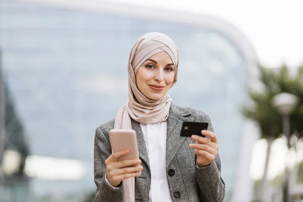 Mujer musulmana en hijab usando teléfono celular con tarjeta bancaria de crédito, haciendo pago en línea — Foto de Stock