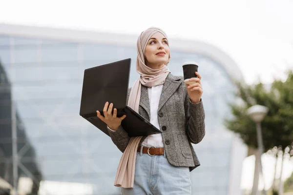 Mulher muçulmana em desgaste casual inteligente, de pé fora do edifício moderno e internet surf no laptop — Fotografia de Stock