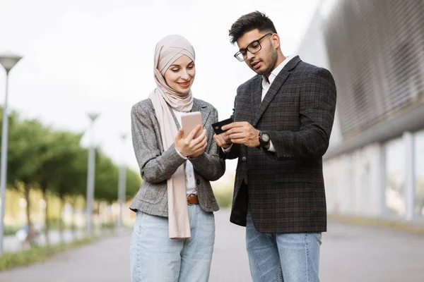 Arabisch-muslimisches Geschäftspaar steht im Freien in der Nähe eines modernen Gebäudes und macht Online-Zahlungen — Stockfoto