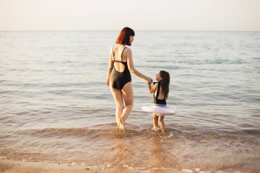 Kızıl saçlı mutlu bir anne ve siyah mayolu küçük bir kız ve kumsalda şişme bir oyuncak.