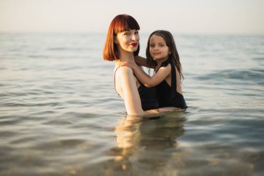 Kızıl saçlı mutlu anne denizde siyah bir mayo giymiş kızını tutuyor..