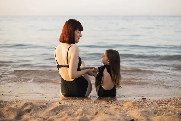 Attraente donna madre con i capelli rossi e la sua piccola figlia carina seduta sulla spiaggia del mare. — Foto Stock
