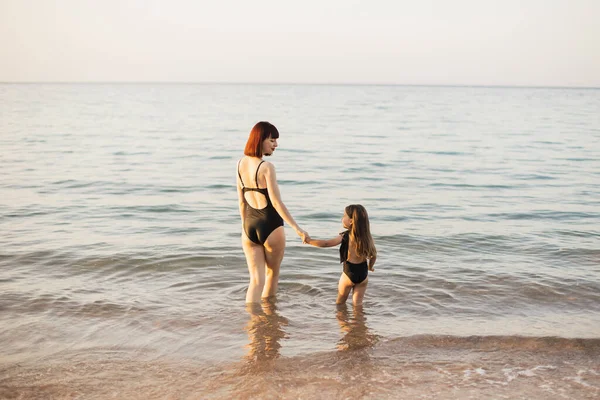 Madri mano condurre la sua bambina in costumi da bagno neri in spiaggia. — Foto Stock