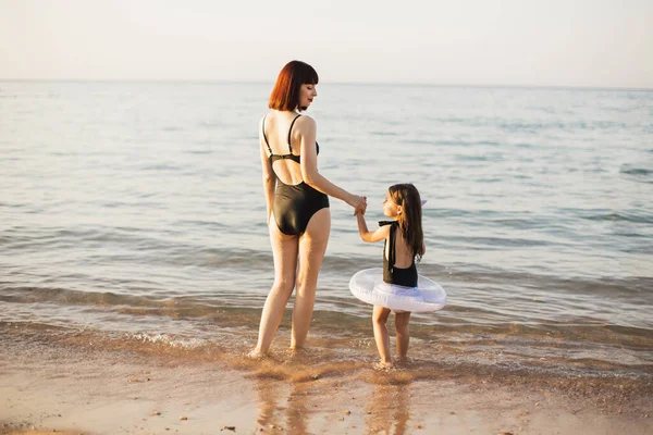 Madre felice con i capelli rossi e figlia giovane in costume da bagno nero e giocattolo gonfiabile sulla spiaggia — Foto Stock