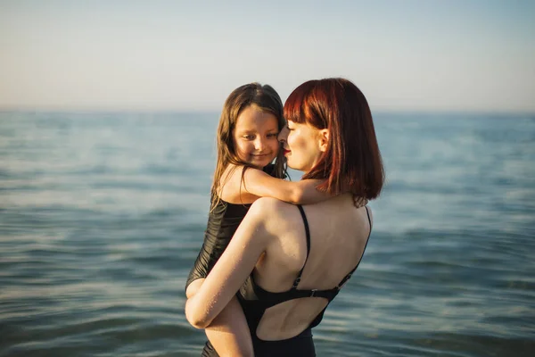 Felice madre in costume da bagno nero con piccola figlia abbraccia le spalle sulla spiaggia. — Foto Stock