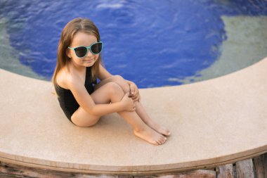 Siyah mayo giymiş şirin bir kız havuzun kenarında yatıyor.