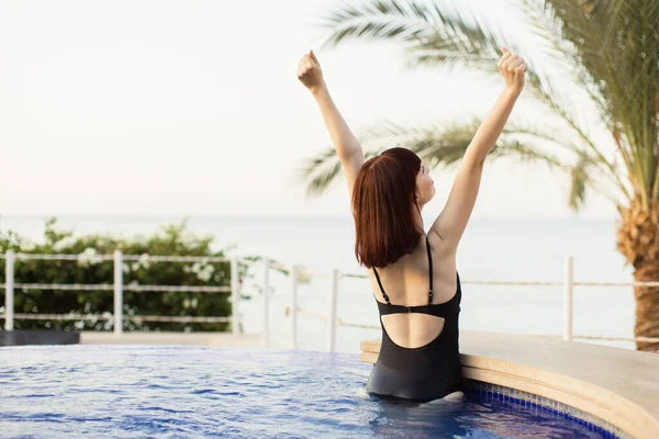 Kaukasische Dame im schwarzen Badeanzug posiert im Pool des Resorts und blickt mit erhobenen Armen in den Himmel — Stockfoto