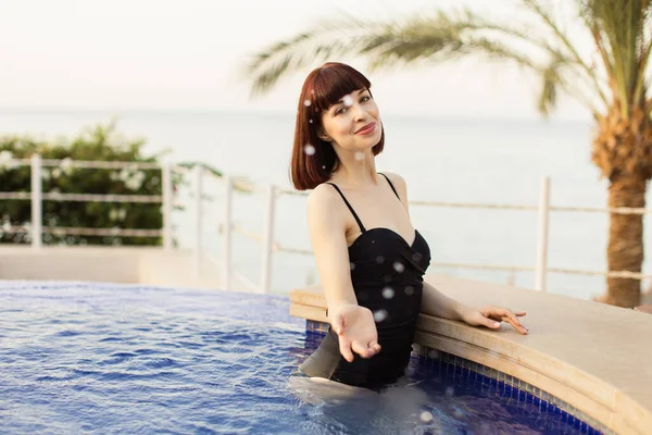 검은 수영복을 입은 매력적 인 젊은 여성, 수영장에서 서 카메라로 물을 튀기는 모습 — 스톡 사진