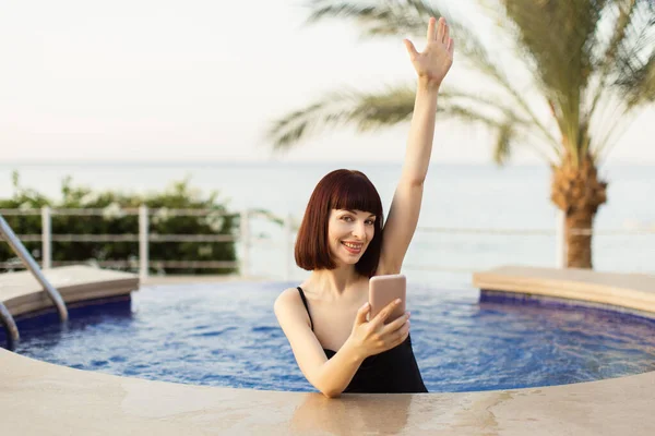 Fröhliches kaukasisches Mädchen im schwarzen Badeanzug, mit Smartphone in der Hand, entspannt sich am sonnigen Tag im Pool — Stockfoto