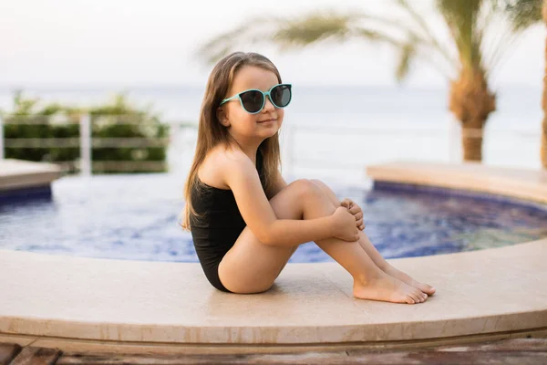 Sonriente linda niña caucásica con gafas de sol azules en la piscina al aire libre en día soleado. — Foto de Stock