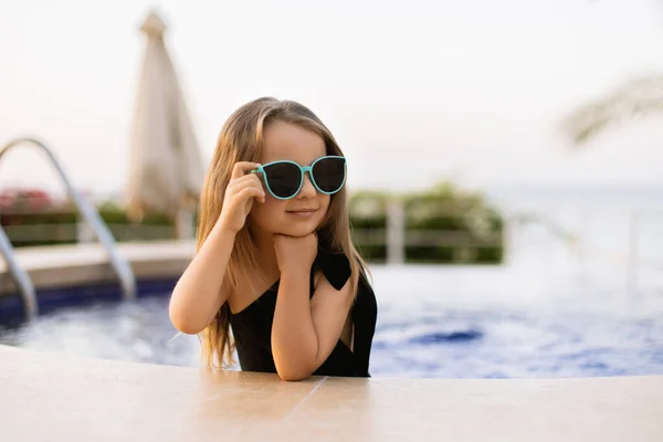 Liebenswertes kleines Mädchen am Pool in Badeanzug und Sonnenbrille, das während der Sommerferien Spaß hat — Stockfoto