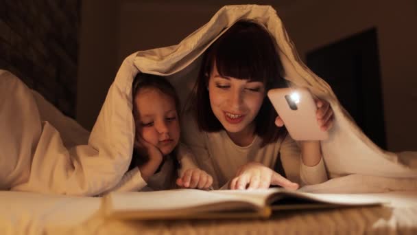 Fiatal szerető anya és gyermek lánya könyvet olvas elemlámpával lefekvés előtt — Stock videók