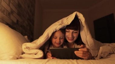 Genç anne, tatlı küçük kızıyla yatakta uzanıyor, tablet bilgisayarına bakıyor ve komik çizgi film izliyor.