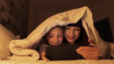 Neşeli anne ve anaokulu kızı, akşam yatak odasında tabletle oynuyorlar.