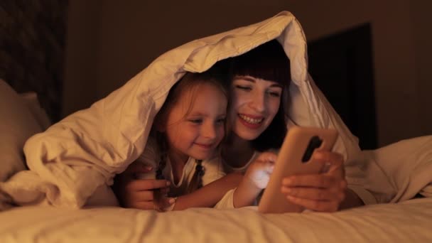 Mama leżąca na łóżku pod kocem z córką, korzystająca z aplikacji mobilnej, spędzająca wieczorny czas — Wideo stockowe