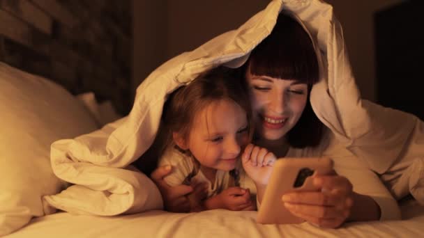 Pretty mama relaks z małą dziewczynką dziecko za pomocą telefonu komórkowego razem, leżąc na łóżku pod kocem — Wideo stockowe