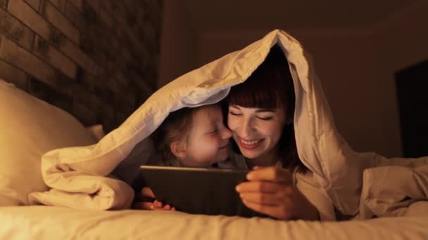 Młoda matka i jej córka leżące na łóżku pod kocem z tabletem, oglądające film — Wideo stockowe