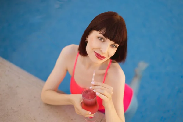 Mujer en traje de baño rosa disfrutando de la natación en la piscina del complejo de lujo en el soleado día de verano con cóctel —  Fotos de Stock