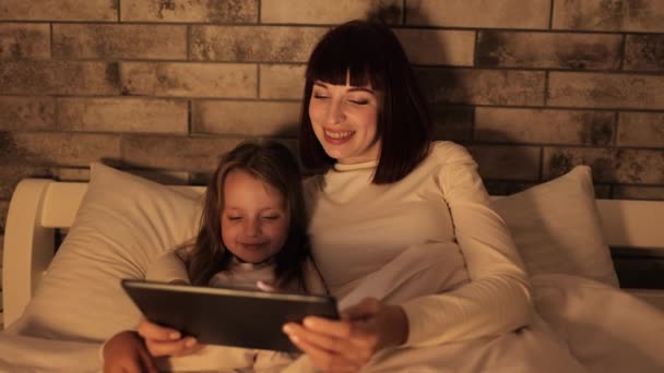 Matka i mała przedszkolanka, leżąca pod kocem na łóżku, oglądająca wideo na tablecie — Wideo stockowe
