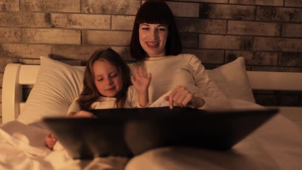 Śliczna uśmiechnięta mama i słodka córeczka oglądają film na laptopie, leżąc w łóżku pod kocem — Wideo stockowe