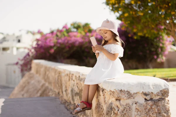 Kleines Mädchen in weißem Kleid und Strohhut schaut sich das Video mit ihrem Smartphone an — Stockfoto