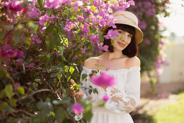 Portrait d'une charmante jeune femme en robe blanche et chapeau de paille debout près d'un arbre rose en fleurs — Photo