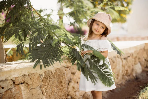 Bonito menina caucasiana em um vestido branco e chapéu de palha fica perto de uma árvore tropical verde — Fotografia de Stock