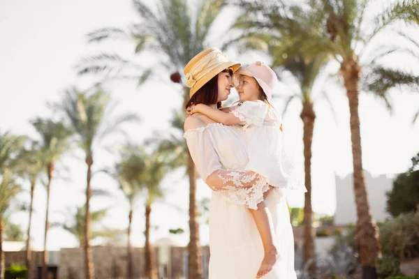 Femme heureuse et sa fille en robes blanches et chapeaux de paille bénéficiant d'une belle vue avec des palmiers — Photo