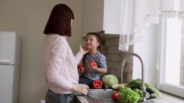 Uma jovem mãe bonita e sua filha pequena bonito lavar legumes sob a torneira com água — Vídeo de Stock