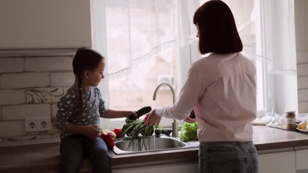 Jovem mãe caucasiana feliz com filha bonita na cozinha lavar legumes na pia — Vídeo de Stock
