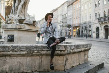 Şehir caddesinde oturan Afrikalı adam cep telefonuyla konuşuyor.