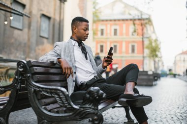 Lüks takım elbiseli Afrikalı bir adam açık havada bankta oturuyor.