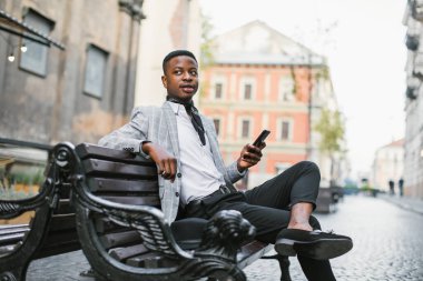 Lüks takım elbiseli Afrikalı bir adam açık havada bankta oturuyor.