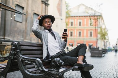 Takım elbiseli ve şapkalı Afrikalı adam akıllı telefonuyla bankta oturuyor.