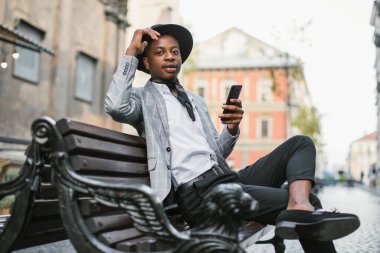 Takım elbiseli ve şapkalı Afrikalı adam akıllı telefonuyla bankta oturuyor.