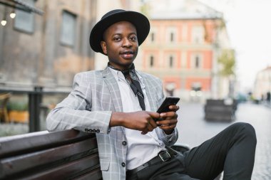 Afrikalı adam bankta dinlenirken akıllı telefondan mesaj atıyor.
