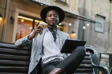 Kahve ve tabletle bankta oturan şık bir Afrikalı.