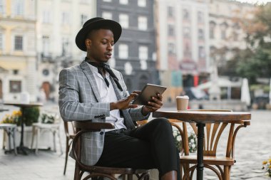 Kafe terasında kahve ve tabletle oturan Afrikalı bir adam.