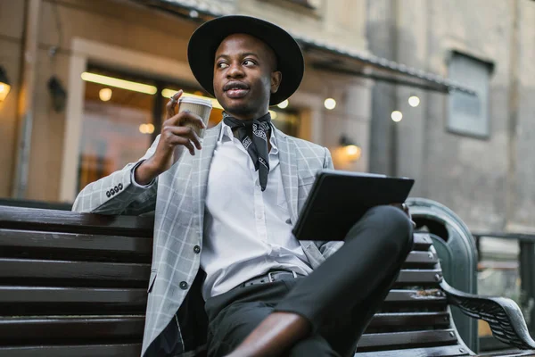 Afrikalı adam kahve içiyor ve bankta tablet kullanıyor.