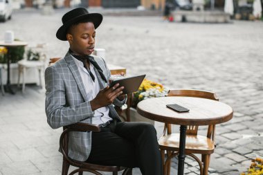 Kafe terasında kahve ve tabletle dinlenen Afrikalı bir adam.