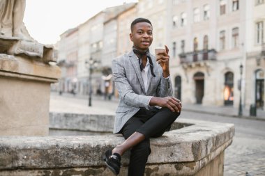 Afrikalı bir adam elinde kahveyle şehir caddesinde oturuyor.