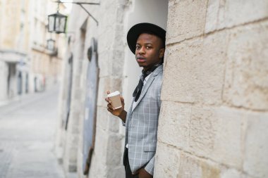 Şapkalı Afrikalı adam elinde kahve fincanıyla sokakta poz veriyor.