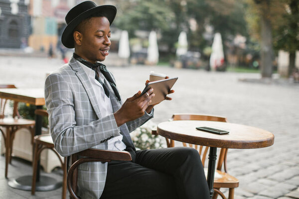 Африканский мужчина отдыхает на террасе кафе с кофе и планшетом