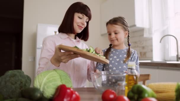 Nette Tochter bereiten Mittagessen mit Mama werfen geschnittene Gurken in Glasschüssel in modernen Küchenraum — Stockvideo