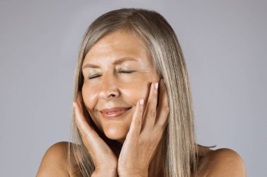 Yaşlı kadın gözlerini kapatıyor ve yanaklarına dokunuyor.